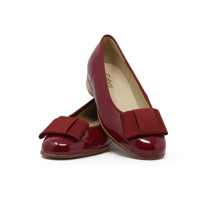 Red leather ballet flats with bow strap and low block heel, angled view.